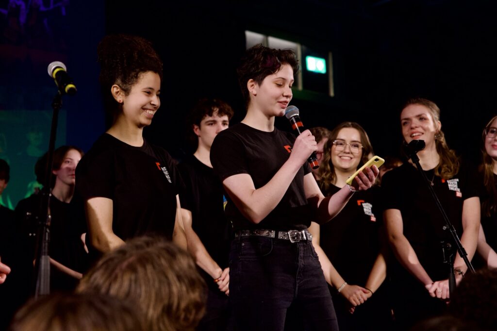 Two young people stand in front of a group of young people, one speaking into a microphone addressing an audience. All are in black BYMT tshirts.